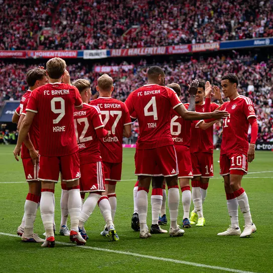 FC Bayern Munich players are gathered in a huddle on the field, wearing their red and white team uniforms. They are celebrating or strategizing together, with visible camaraderie among the team members. A packed stadium is visible in the background.