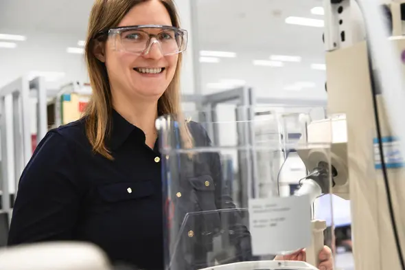 A female scientist wearing safety glasses smiles while standing near laboratory equipment. She is in a well-lit lab environment, with various scientific tools and equipment in the background.