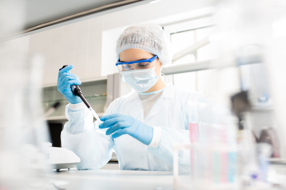 A laboratory technician in protective gear, including a mask, goggles, and gloves, is using a pipette to transfer liquids in a lab setting. The technician is focused on their task, with various lab equipment in the background.