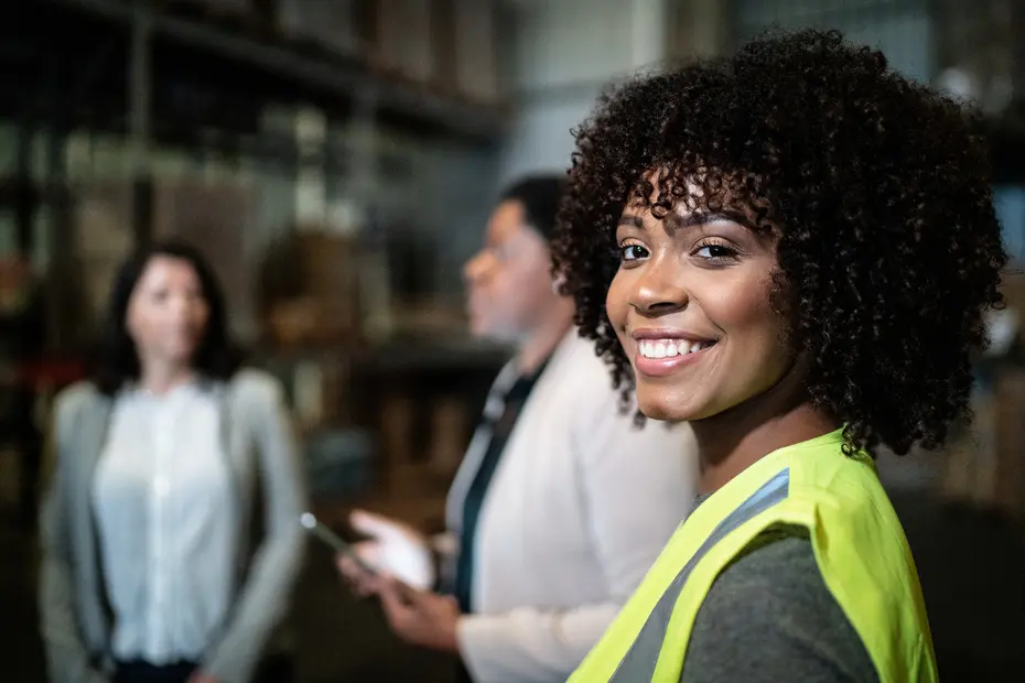 A smiling woman with curly hair, wearing a bright yellow safety vest, stands in a warehouse setting. Behind her, two other women are engaged in conversation, creating a collaborative atmosphere in the industrial environment.