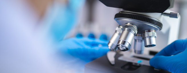 A close-up view of a microscope with a person's gloved hand adjusting the slide. The background is blurred, suggesting a laboratory setting.