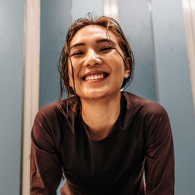 A smiling young woman with wet hair leans forward, exuding happiness and vitality. She is wearing a dark, fitted shirt, and the background features light blue walls. The image captures a moment of joy and energy, likely after physical activity.