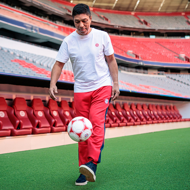 An individual in a white t-shirt and red sweatpants is skillfully juggling a soccer ball on the field of a stadium. The empty stands are visible in the background, highlighting the vibrant seats. The atmosphere suggests a focus on sports and athleticism.