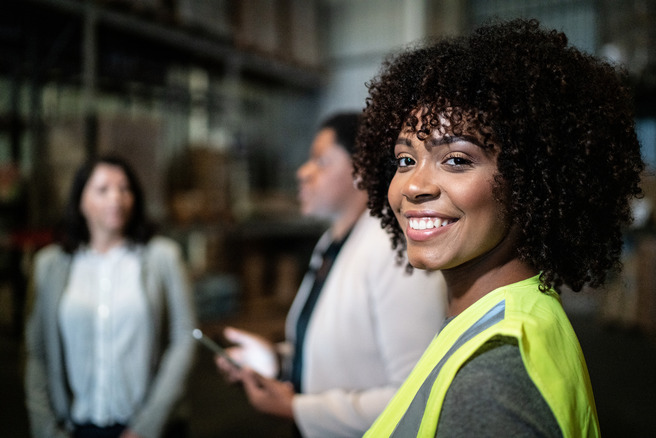 A smiling woman with curly hair, wearing a bright yellow safety vest, stands in a warehouse setting. Behind her, two other women are engaged in conversation, creating a collaborative atmosphere in the industrial environment.