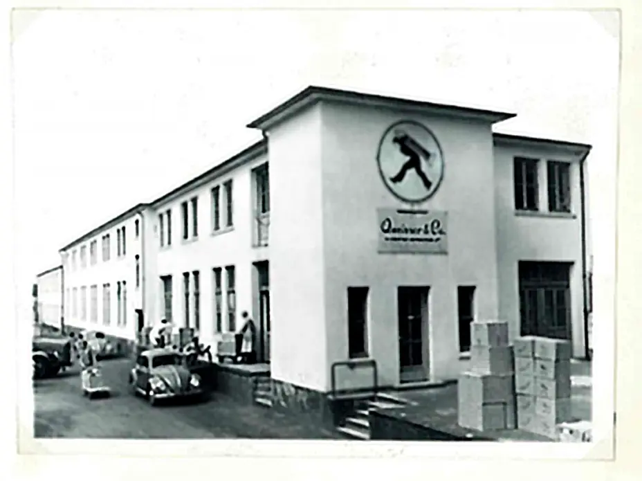 A historic black and white photo of a rectangular building with large windows and a logo featuring a walking figure. Boxes are stacked near the entrance, and a few people and vintage cars are visible in the foreground. The environment suggests an industrial or commercial setting.