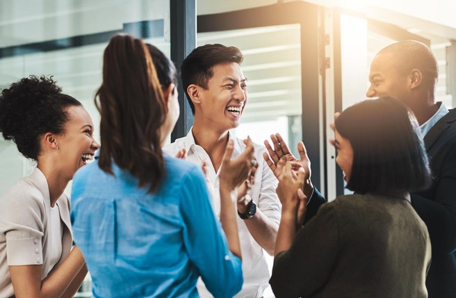 A diverse group of five people, two men and three women, are joyfully interacting in an office setting. They are smiling, laughing, and clapping, creating a positive and lively atmosphere. The scene conveys a sense of teamwork and celebration.