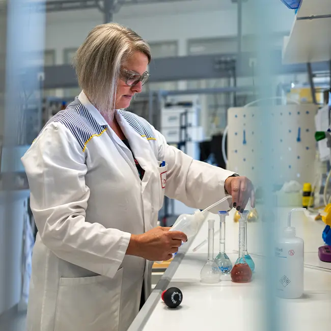 Eine Laborangestellte arbeitet mit Erlenmeyerkolben und Pipetten. Im Hintergrund ist weitere Laborausruestung sichtbar.