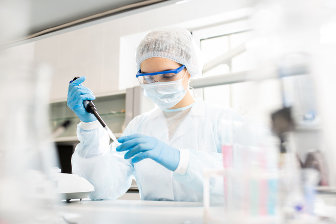 A laboratory technician in protective gear, including a mask, goggles, and gloves, is using a pipette to transfer liquids in a lab setting. The technician is focused on their task, with various lab equipment in the background.