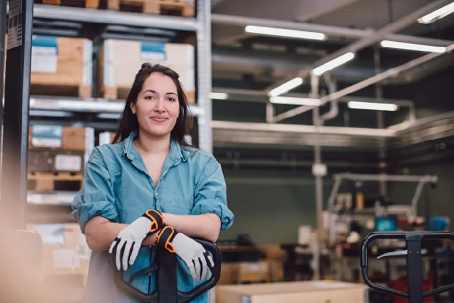 A woman wearing gloves stands confidently next to a pallet jack in a warehouse. She has long hair and wears a denim shirt, with shelves of stacked boxes visible in the background, indicating a industrial setting. 