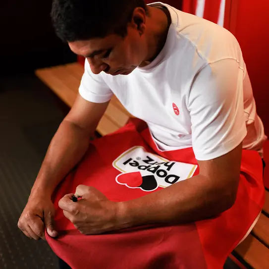 A man in a white t-shirt sits on a bench in a locker room, focused on adjusting a red towel with a graphic design. He appears engaged and calm, surrounded by a simple backdrop of red and wooden elements.