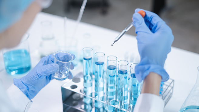 A laboratory scene featuring a scientist in gloves, holding a pipette above a rack of test tubes filled with blue liquid. In the other hand, the scientist holds a smaller container of blue liquid, suggesting a process of measurement or mixing.