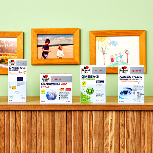 A wooden shelf displays several health product boxes, including Omega-3 liquid, Magnesium 400 Citrate, and Omega-3 plant-based. The backdrop features a green wall with three framed photos and a colorful drawing by children, creating a warm, homey atmosphere.