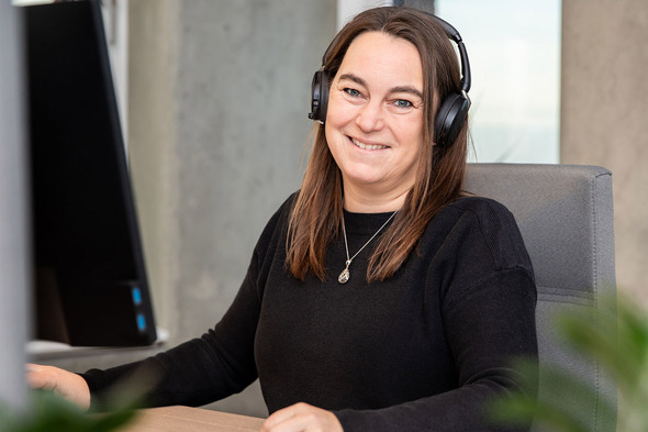 A woman with long dark hair is sitting at a desk, smiling while wearing headphones. She is facing a computer monitor and appears engaged and friendly in her work environment. Soft green plants are slightly visible in the foreground.