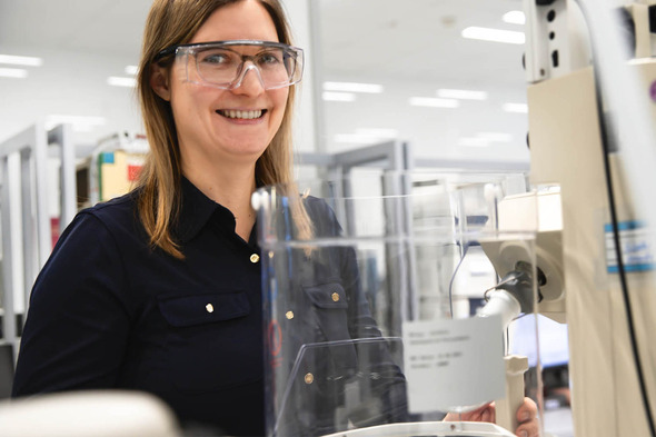 A female scientist wearing safety glasses smiles while standing near laboratory equipment. She is in a well-lit lab environment, with various scientific tools and equipment in the background.