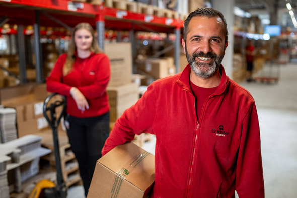 Two warehouse workers are in the foreground, one smiling and holding a box, while the other stands behind with a pallet jack. Both are wearing red shirts, and the background features shelves stacked with boxes, suggesting a busy work environment.